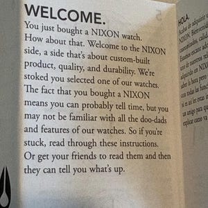 May include: A folded paper with the word "WELCOME" in large, bold letters. The text welcomes the buyer of a NIXON watch and provides instructions. The paper is off-white with black text.