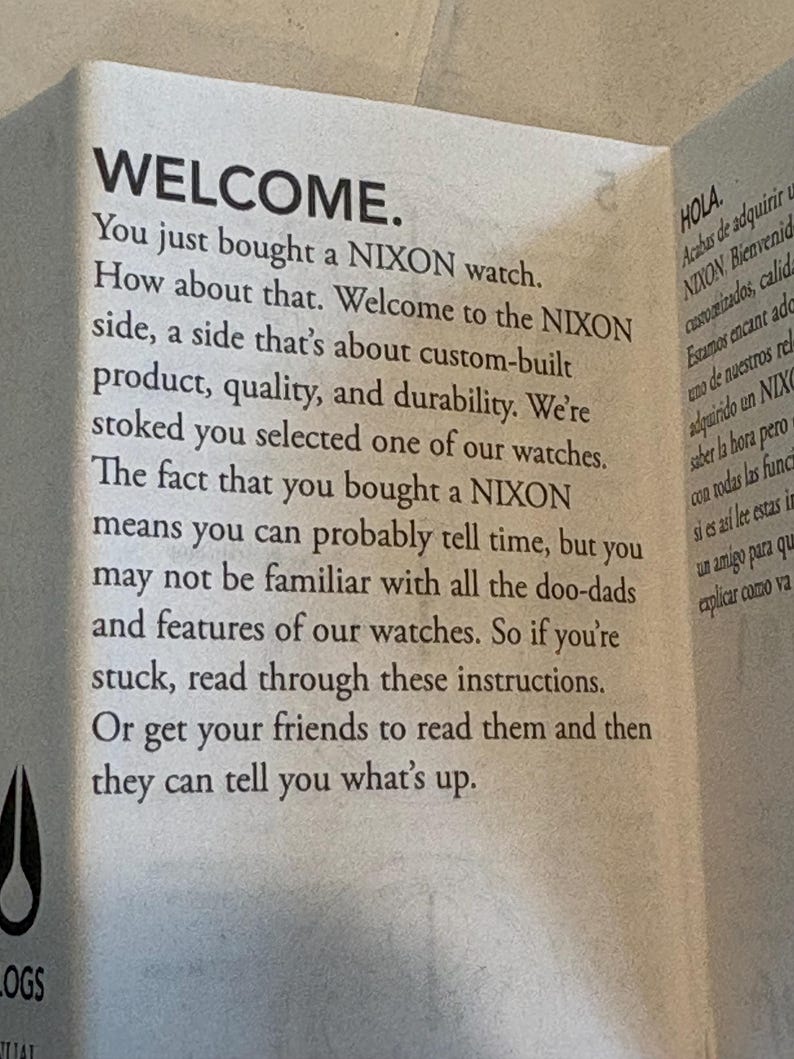 May include: A folded paper with the word "WELCOME" in large, bold letters. The text welcomes the buyer of a NIXON watch and provides instructions. The paper is off-white with black text.