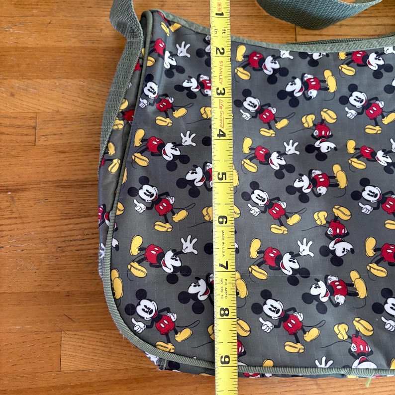 Puede incluir: Un bolso de mensajero gris con un patr&oacute;n repetitivo de Mickey Mouse con pantalones cortos rojos y zapatos amarillos. Una cinta m&eacute;trica amarilla est&aacute; colocada verticalmente sobre el bolso, mostrando una longitud de unos 23 cm de arriba a abajo.