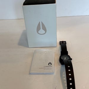 May include: A black analog watch with a black band and a round black watch face. The watch is next to a white box with a black top and a logo. An instruction manual is also present.