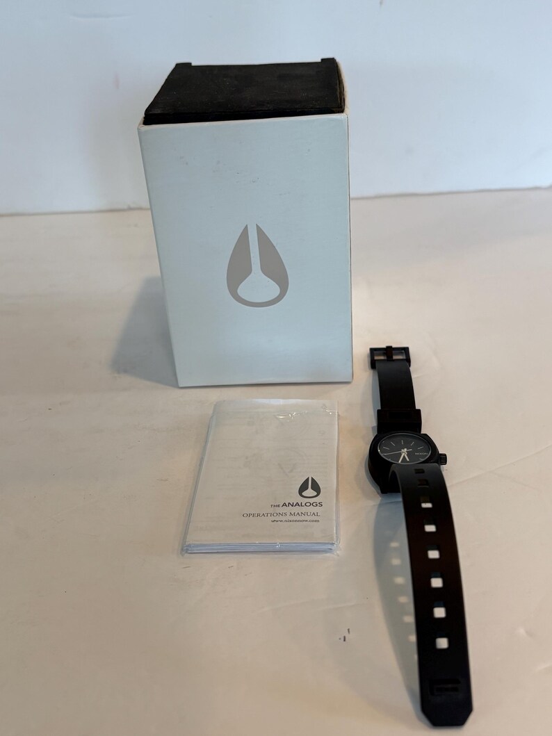 May include: A black analog watch with a black band and a round black watch face. The watch is next to a white box with a black top and a logo. An instruction manual is also present.