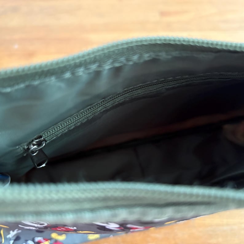 Puede incluir: Un estuche con cremallera de color verde oliva con un forro interior a juego. El interior incluye un bolsillo con cremallera con un tirador de metal plateado. La tela exterior muestra un patr&oacute;n repetitivo de personajes de dibujos animados.