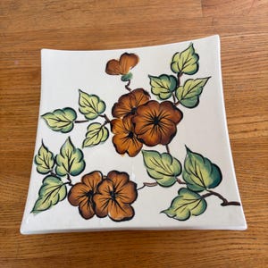 Könnte beinhalten: Quadratischer, cremefarbener Keramikteller mit floralem Design. Die Blumen sind braun mit orangefarbenen Zentren, und die Blätter sind grün und gelb. Der Teller hat leicht abgerundete Ecken und steht auf einer Holzoberfläche.