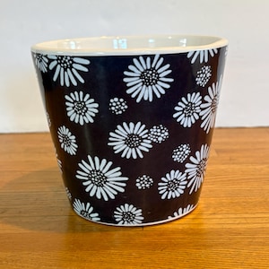 May include: A tapered ceramic planter featuring a dark navy blue background with a repeating pattern of white daisy-like flowers and small white dots. The interior of the vessel is white, and the rim has a smooth, rounded white edge.
