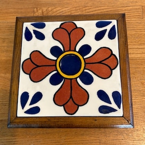May include: A square ceramic tile set within a dark brown wooden frame. The tile features a white background with a central dark blue circle surrounded by a yellow ring, four large rust-colored petal shapes, and four dark blue petal accents.