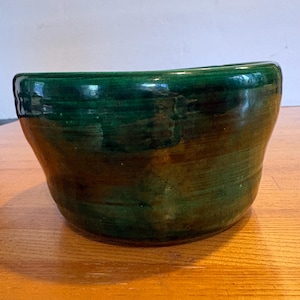 May include: A handcrafted, green ceramic bowl with a glossy finish. The bowl has a wide, open top and a rounded shape. The exterior features variations in the green glaze, creating a textured appearance. This decorative bowl is suitable for serving or display.