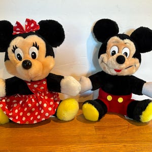 May include: Two plush Disney character dolls, Mickey Mouse and Minnie Mouse, are seated side-by-side. Minnie wears a red polka-dot dress and bow. Mickey has red shorts with yellow buttons. Both have black ears, black bodies, and yellow feet.