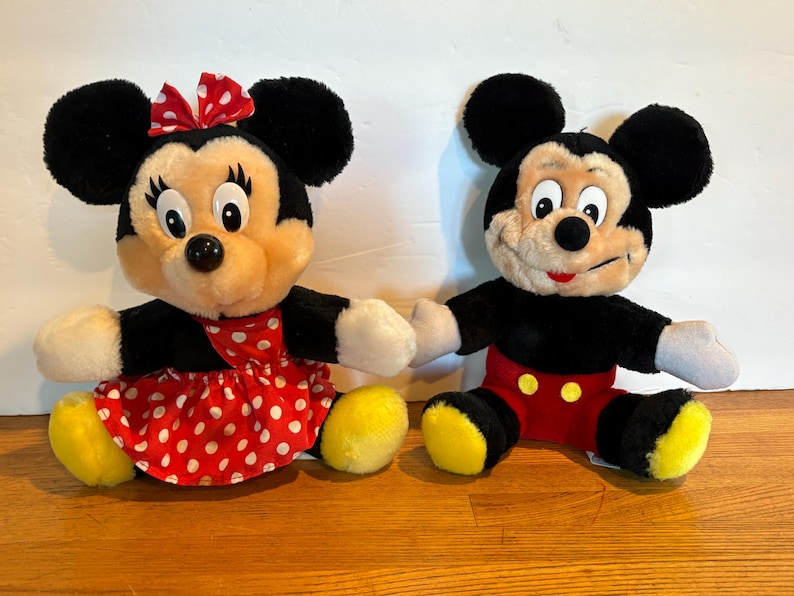 May include: Two plush Disney character dolls, Mickey Mouse and Minnie Mouse, are seated side-by-side. Minnie wears a red polka-dot dress and bow. Mickey has red shorts with yellow buttons. Both have black ears, black bodies, and yellow feet.