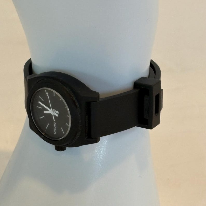 May include: A black Nixon watch with a round face and a black band. The watch has white hands and markers against a black background. The watch is displayed on a white surface.