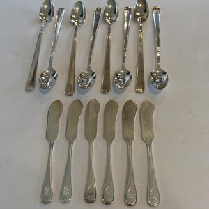 Könnte beinhalten: Eine Sammlung von silberfarbenem Besteck, darunter Löffel und Buttermesser. Die Löffel haben längliche Griffe und abgerundete Laffen. Die Buttermesser haben gebogene Klingen und ein dekoratives Emblem am Griff. Das Besteck ist auf einer hellen Oberfläche angeordnet.