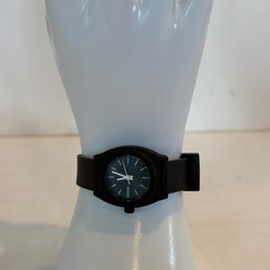 May include: A black Nixon watch with a round face and a black leather band. The watch has a dark face with white hands and markers. The watch is displayed on a white mannequin hand.