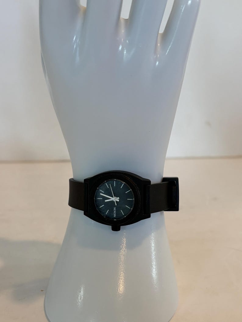 May include: A black Nixon watch with a round face and a black leather band. The watch has a dark face with white hands and markers. The watch is displayed on a white mannequin hand.