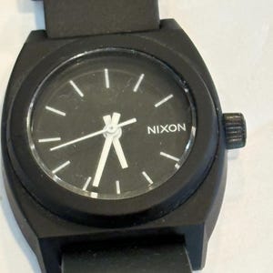 May include: Black Nixon watch with a round black face and white hour markers. The watch has a black band and a black case. The word "NIXON" is printed in white on the watch face.