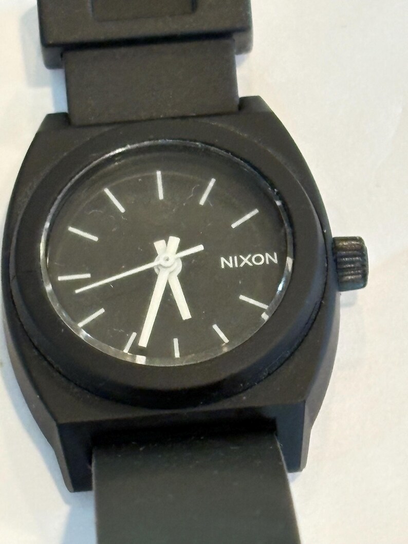 May include: Black Nixon watch with a round black face and white hour markers. The watch has a black band and a black case. The word "NIXON" is printed in white on the watch face.