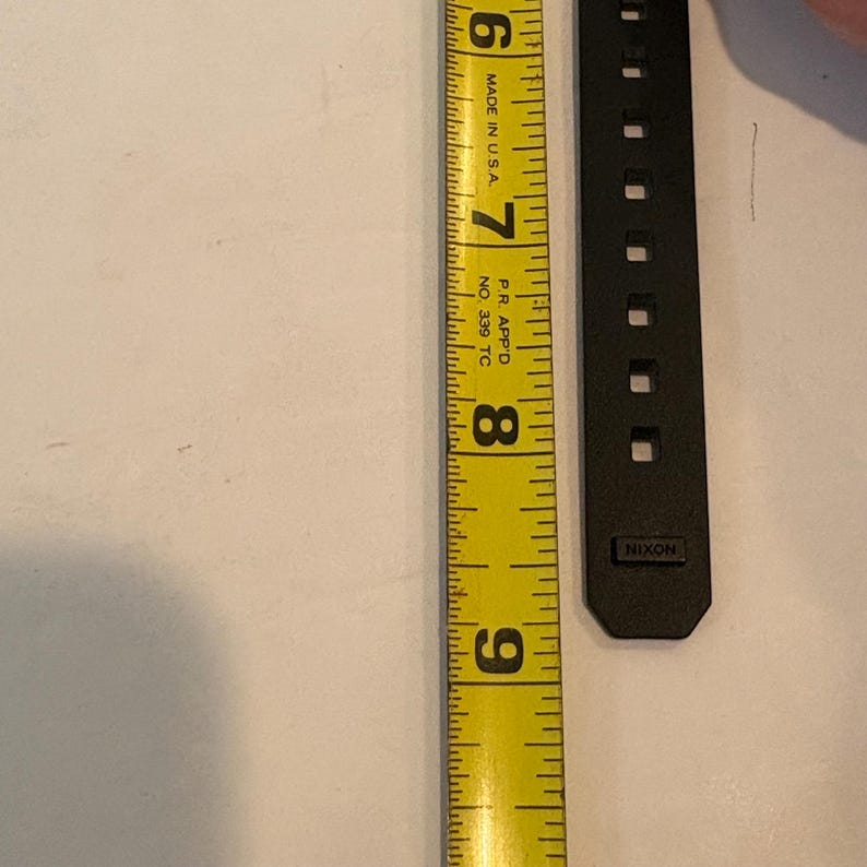 May include: A black watch band next to a yellow measuring tape. The tape shows measurements from 6 to 9 inches. The watch band has the brand name "Nixon" printed on it.