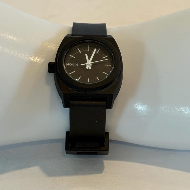 May include: A black Nixon watch with a dark face and white hands and markers. The watch has a black band and a black case. The watch is displayed on a white surface.