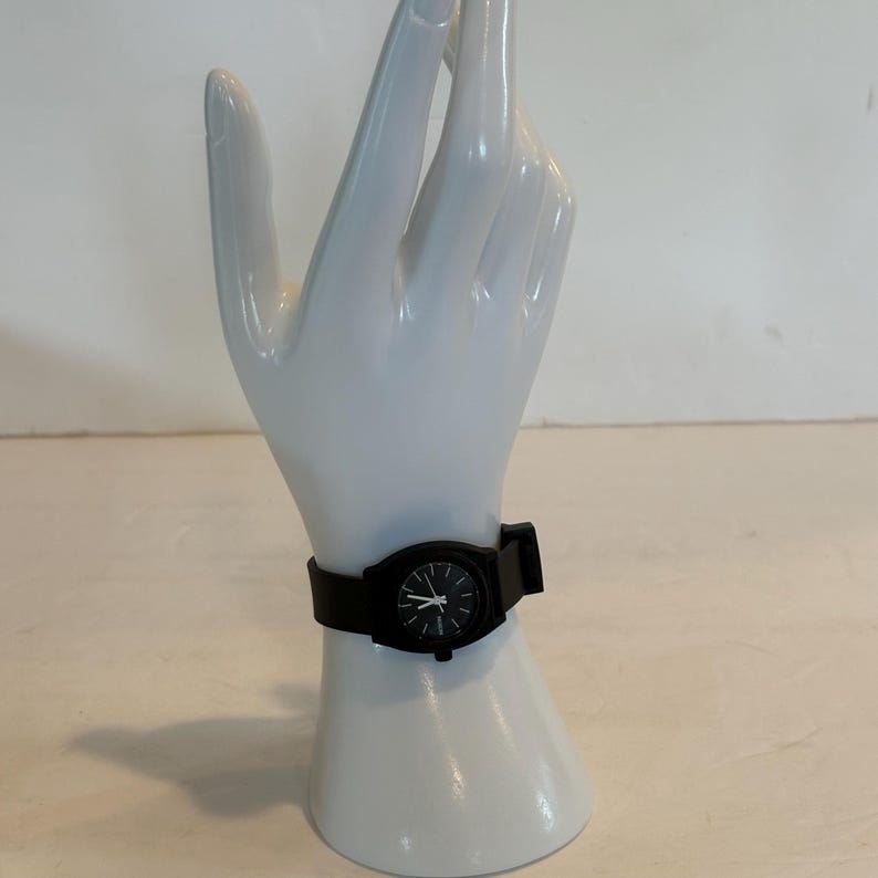 May include: A black wristwatch with a black band. The watch face is black with white hands and markers. The watch is displayed on a white hand-shaped display.