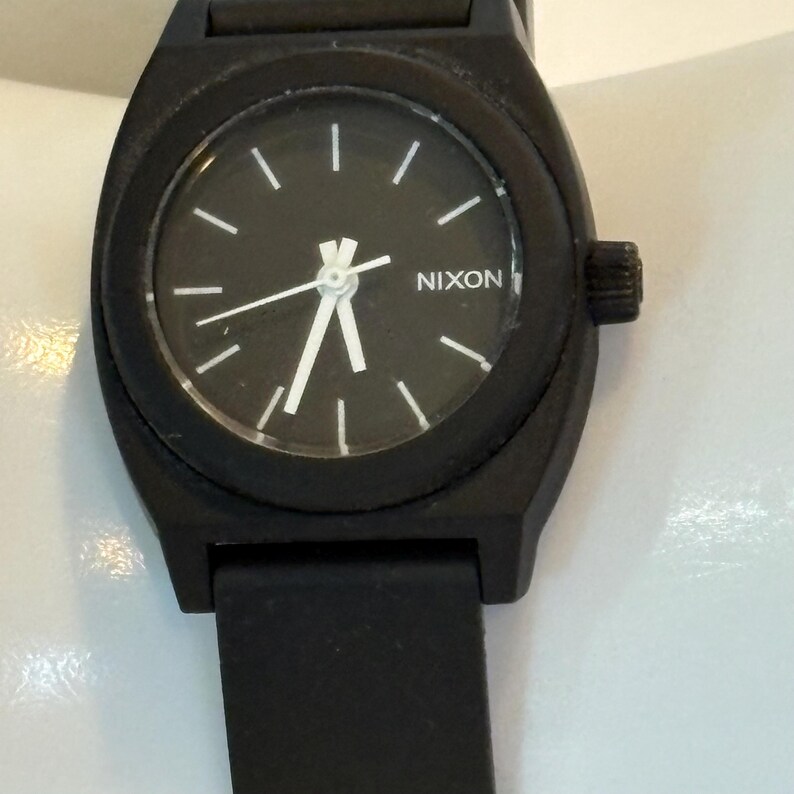 May include: A black Nixon watch with a round face and a black band. The watch face is black with white hour markers and hands. The brand name "NIXON" is printed in white.