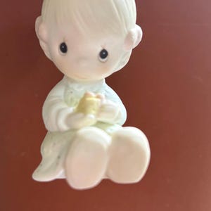 Puede incluir: Una figura de cerámica de un niño sentado con tez clara, vestido con un atuendo azul claro y blanco. El niño sostiene un objeto amarillo. La figura está suspendida por una cadena dorada.