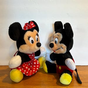 May include: Two plush Disney characters, Mickey and Minnie Mouse, are displayed. Minnie Mouse wears a red polka-dot dress and bow. Mickey Mouse is in red shorts. Both have black fur, white gloves, and yellow feet.