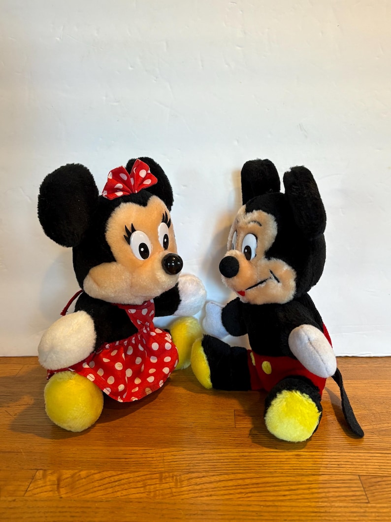 May include: Two plush Disney characters, Mickey and Minnie Mouse, are displayed. Minnie Mouse wears a red polka-dot dress and bow. Mickey Mouse is in red shorts. Both have black fur, white gloves, and yellow feet.