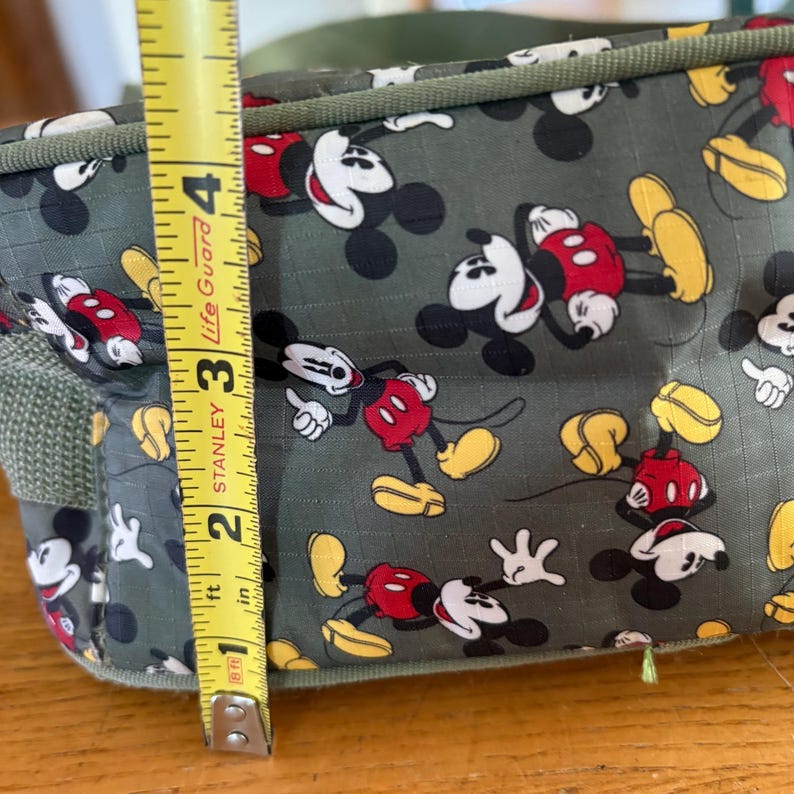 Puede incluir: Una bolsa de tela verde oliva con un patr&oacute;n repetitivo de Mickey Mouse con pantalones cortos rojos y zapatos amarillos. Una cinta m&eacute;trica amarilla Stanley est&aacute; colocada verticalmente en el lateral, mostrando una altura de unos 10 cm.