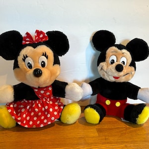 May include: Two plush Disney character dolls, Mickey and Minnie Mouse, sit side-by-side. Minnie wears a red polka-dot dress and bow. Mickey wears red shorts. Both have black fur, yellow shoes, and white gloves.