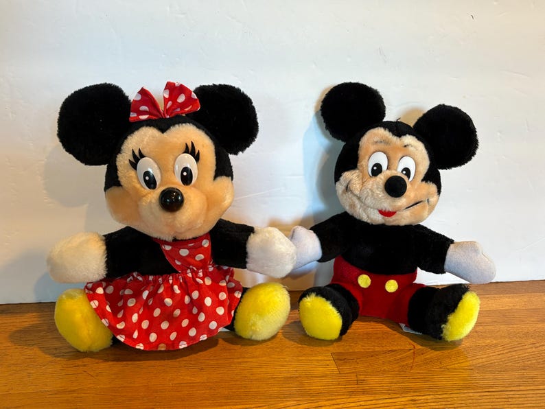 May include: Two plush Disney character dolls, Mickey and Minnie Mouse, sit side-by-side. Minnie wears a red polka-dot dress and bow. Mickey wears red shorts. Both have black fur, yellow shoes, and white gloves.