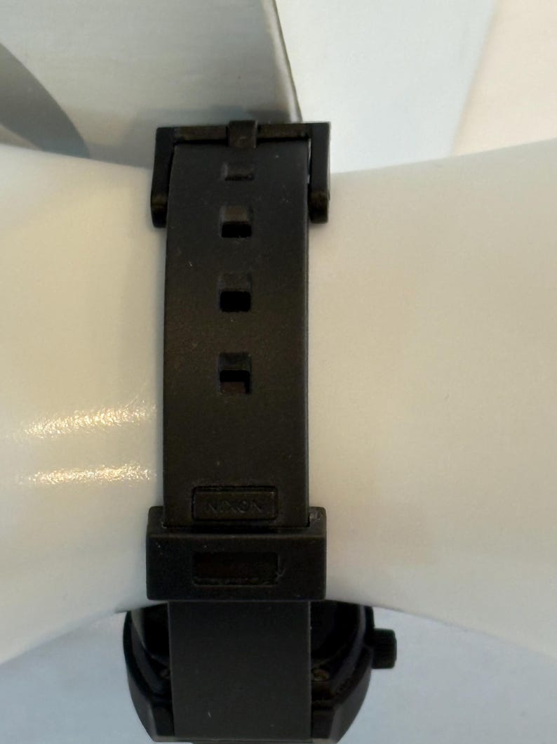 May include: Black watch with a black band. The band is made of a flexible material and has a buckle closure. The watch face is visible at the bottom of the image.