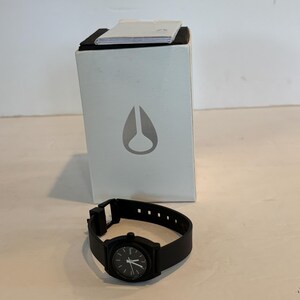 May include: A black wristwatch with a black band and a black watch face. The watch is next to a white box with a gray logo. The watch band has multiple holes for adjustment.