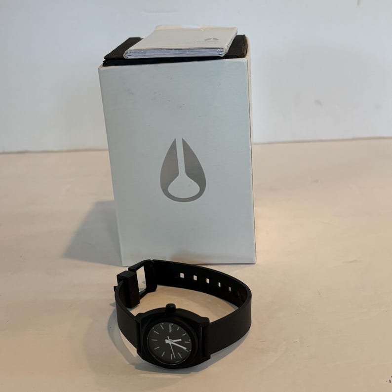 May include: A black wristwatch with a black band and a black watch face. The watch is next to a white box with a gray logo. The watch band has multiple holes for adjustment.
