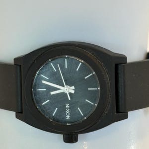 May include: A black Nixon watch with a dark gray face and white hour markers. The watch has a black band and the word "NIXON" is printed on the watch face. The watch is a minimalist design.
