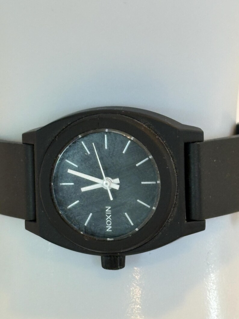 May include: A black Nixon watch with a dark gray face and white hour markers. The watch has a black band and the word "NIXON" is printed on the watch face. The watch is a minimalist design.