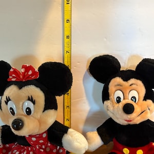 May include: Two plush Disney character dolls: Minnie Mouse and Mickey Mouse. Minnie wears a red polka-dot dress and bow. Mickey has red shorts. A yellow measuring tape shows the dolls are approximately 11 inches tall.