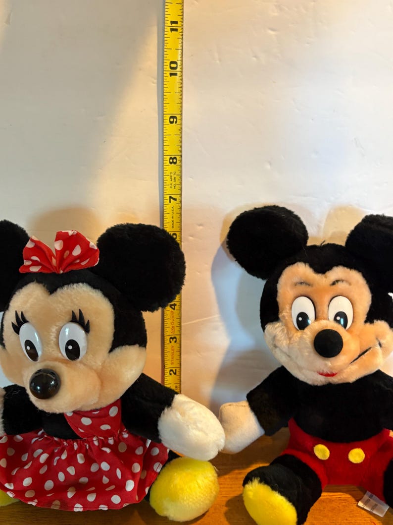 May include: Two plush Disney character dolls: Minnie Mouse and Mickey Mouse. Minnie wears a red polka-dot dress and bow. Mickey has red shorts. A yellow measuring tape shows the dolls are approximately 11 inches tall.