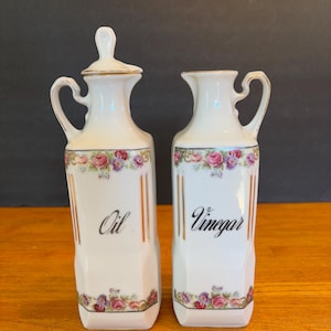 May include: A pair of white ceramic oil and vinegar cruets. The cruets have a floral design with pink and purple roses. The word "Oil" is written on one cruet, and "Vinegar" on the other. Both have gold trim.