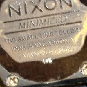 May include: Close-up of a watch face with the brand name "NIXON" in embossed lettering. Below, the text reads "MINIMIZED", "THE SMALL TIME TELLERS", "100M POR CARBONA", and "JAPAN MOVT". The number "14E" is also visible.