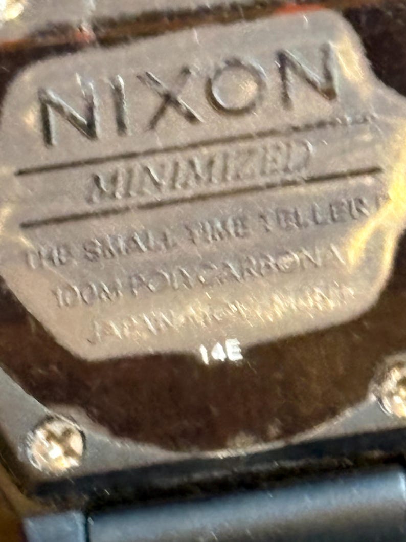 May include: Close-up of a watch face with the brand name "NIXON" in embossed lettering. Below, the text reads "MINIMIZED", "THE SMALL TIME TELLERS", "100M POR CARBONA", and "JAPAN MOVT". The number "14E" is also visible.