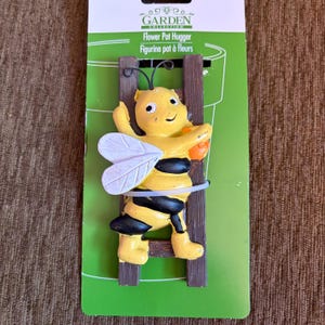 May include: A yellow and black bee figurine climbing a brown ladder, designed as a flower pot hugger. The bee has white wings and is attached to a green card with the text "GARDEN COLLECTION" and "Flower Pot Hugger".