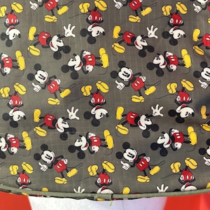Puede incluir: Una tela gris con un patr&oacute;n repetitivo de personajes de Mickey Mouse. Cada figura aparece en una pose cl&aacute;sica con orejas negras, cara blanca, pantalones cortos rojos y zapatos amarillos. El material tiene una sutil textura de cuadr&iacute;cula.
