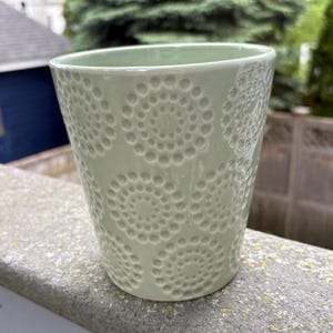 May include: A light green ceramic cup with a textured pattern of concentric circles. The cup is cylindrical with a slightly flared top. The design is embossed on the surface, creating a tactile and visual appeal. The cup is sitting on a concrete surface.