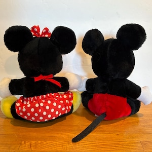 May include: Two plush toys of Mickey and Minnie Mouse, viewed from the back. Mickey has a red shorts and a black tail. Minnie wears a red skirt with white polka dots and a red bow. Both have black ears and white gloves.