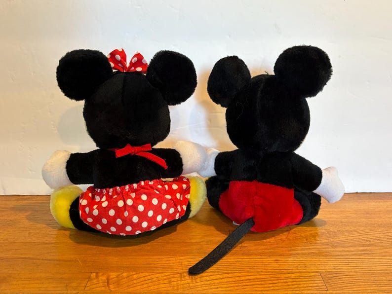 May include: Two plush toys of Mickey and Minnie Mouse, viewed from the back. Mickey has a red shorts and a black tail. Minnie wears a red skirt with white polka dots and a red bow. Both have black ears and white gloves.