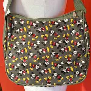 Puede incluir: Un bolso bandolera de color verde oliva con un patr&oacute;n repetitivo de personajes de Mickey Mouse. El bolso cuenta con un cierre de cremallera superior con un tirador de metal plateado y una correa de hombro de tela verde oliva ajustable.