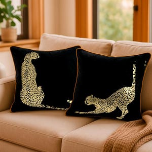 May include: Two black cushions featuring gold cheetah designs. One cheetah is sitting, the other stretching. The cushions have gold piping and are placed on a beige sofa.