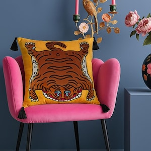 Puede incluir: Un sillón de terciopelo rosa con un cojín amarillo con un diseño de tigre marrón. El cojín tiene borlas en las esquinas. El sillón está frente a una pared azul con una escultura de pájaro dorada y un jarrón con flores rosas.