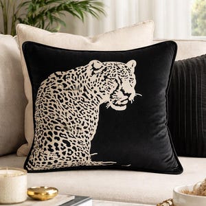 May include: Black square cushion featuring a cream leopard design. The leopard's head and upper body are depicted. The cushion is placed on a cream sofa, with other cushions and decorative items visible in the background.