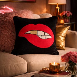 Op de afbeelding: Zwart vierkant decoratief kussen met een groot rood lippenontwerp, witte tanden en een gouden tand. Het kussen ligt op een beige bank, samen met andere decoratieve kussens en een brandende kaars.