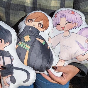Może przedstawiać: Trzy pluszowe poduszki z postaciami w stylu anime. Poduszki są białe z kolorowymi ilustracjami postaci. Jedna postać ma czarne włosy i koci ogon, druga ma brązowe włosy i nosi czarny i zielony strój, a trzecia ma fioletowe włosy i nosi białą koszulę.
