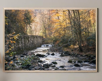 May include: A scenic photograph of a stream flowing through a forest in autumn. The stream is surrounded by colorful trees with yellow, orange, and red leaves. A stone wall runs along the edge of the stream.